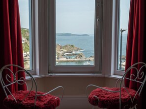 A room with a large window overlooking a coastal view, featuring two metal chairs with red cushions. - Smugglers View by Interhome (Mevagissey)