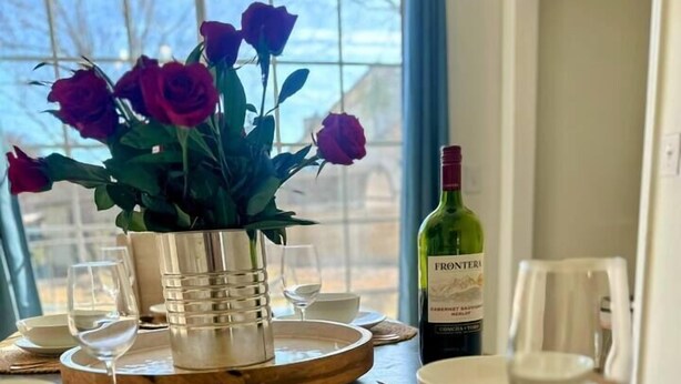 A dining table set with plates, glasses, and a bottle of wine, with a vase of flowers and a window view. - Summerville’s Charm House (Summerville)
