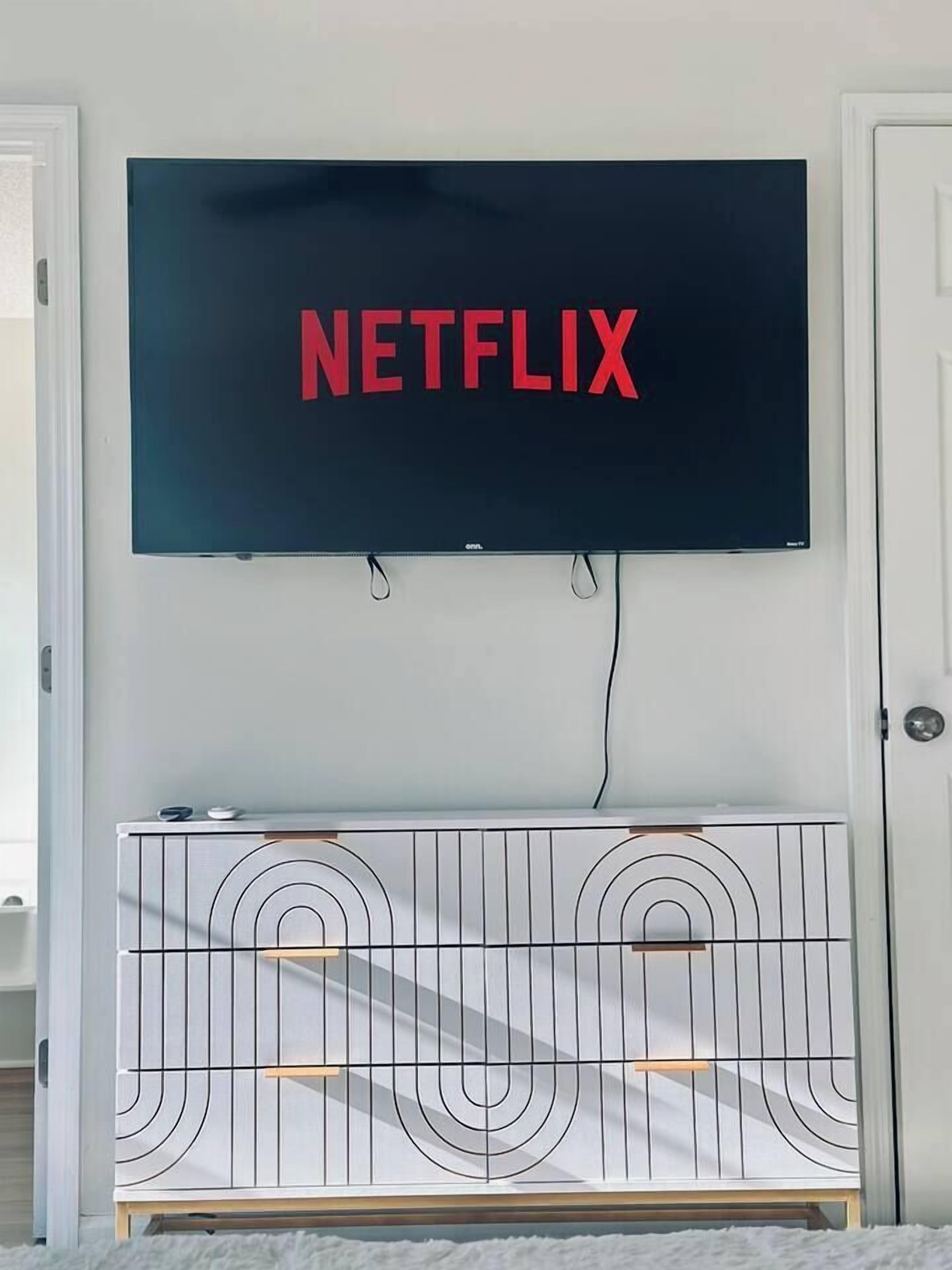 Ein an der Wand befestigter Fernseher mit Netflix-Logo, eine weiße Konsole darunter und eine Tür zur rechten Seite.