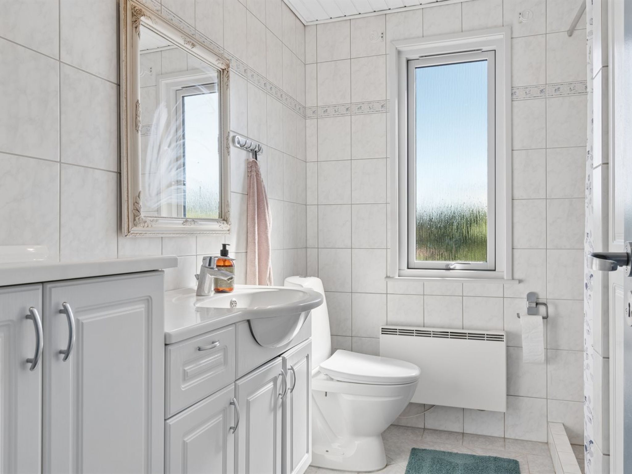 A bathroom with a white toilet, a sink with a soap dispenser, a mirror, and a window with a view of greenery.