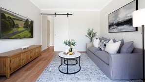 Ein modernes Wohnzimmer mit einer Holzkommode, einem grauen Sofa, einem runden Couchtisch mit einer Vase und Gläsern sowie einem großen Gemälde an der Wand.