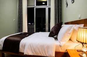 A hotel room with a bed, a nightstand, a lamp, a window with curtains, and a view of the city at night. - KIGALI GOLDEN VILLA RESORTS WITH POOL (Kigali)
