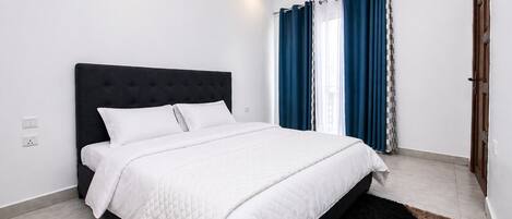 A bedroom with a bed, white bedding, a dark headboard, a black rug, and blue curtains.