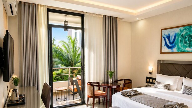 A hotel room with a bed, a desk, a chair, a small table, and a balcony with a view of palm trees. - KIGALI GOLDEN VILLA RESORTS WITH POOL (Kigali)