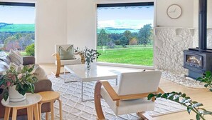 Ett modernt vardagsrum med en stenbeklädd öppen spis, stora fönster med utsikt över ett grönskande landskap, en matplats med bord och stolar samt en soffa.