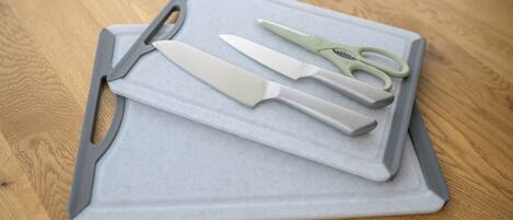 A cutting board with a knife, scissors, and a peeler on a wooden surface.