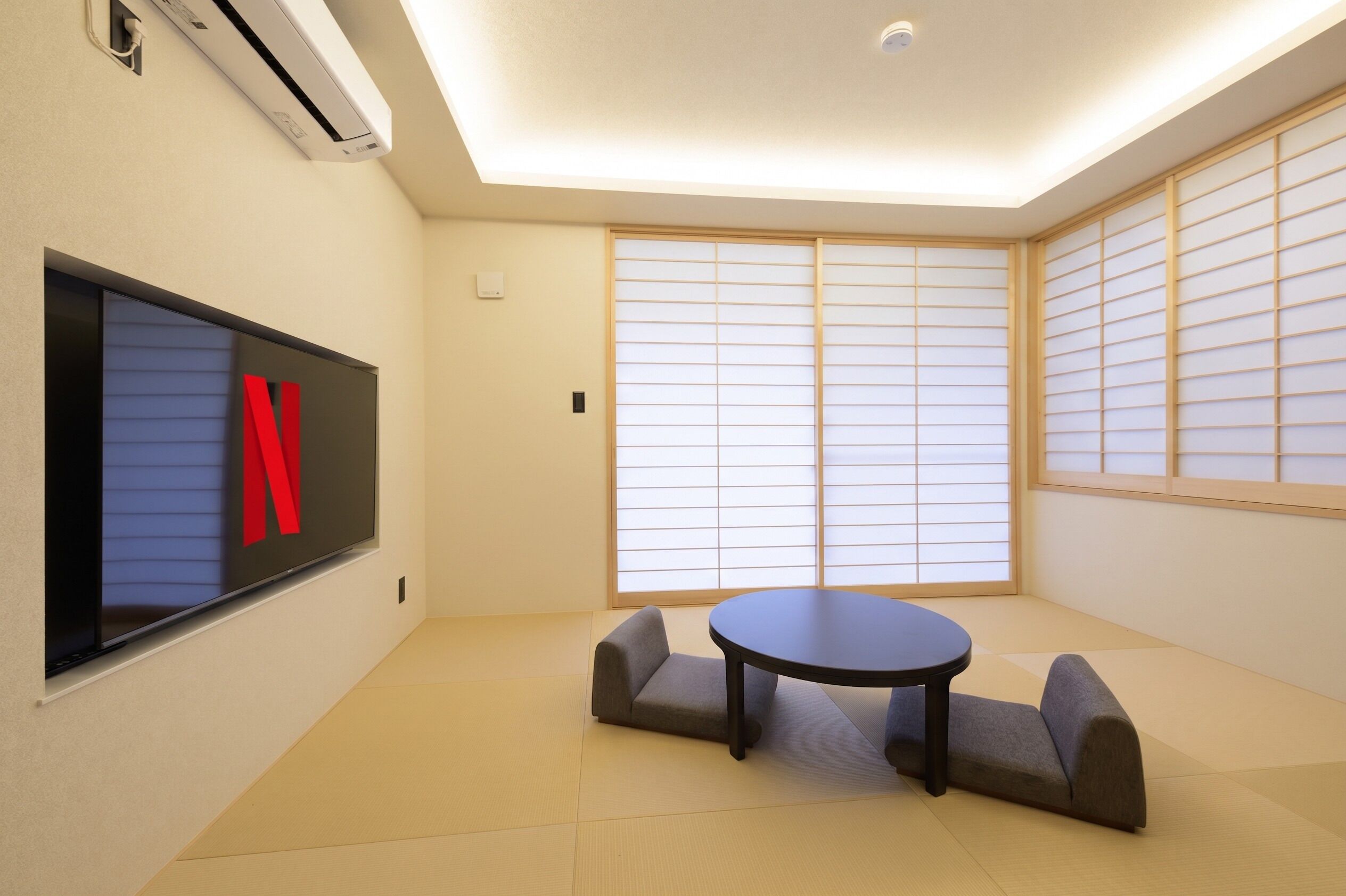 A room with tatami flooring, a round table, two cushioned seating, a wall-mounted TV, and shoji screens.