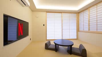 Ein Zimmer mit Tatami-Boden, einem runden Tisch, zwei gepolsterten Sitzgelegenheiten, einem Wand-Fernseher und Shoji-Schirmen.