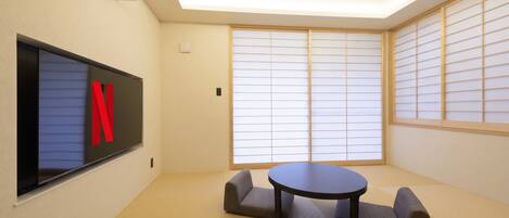 A room with tatami flooring, a round table, two cushioned seating, a wall-mounted TV, and shoji screens.