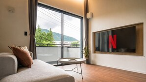 Una sala de estar moderna con un sofá, una mesita redonda, una planta en maceta y un amplio ventanal con vistas a zonas verdes y colinas.