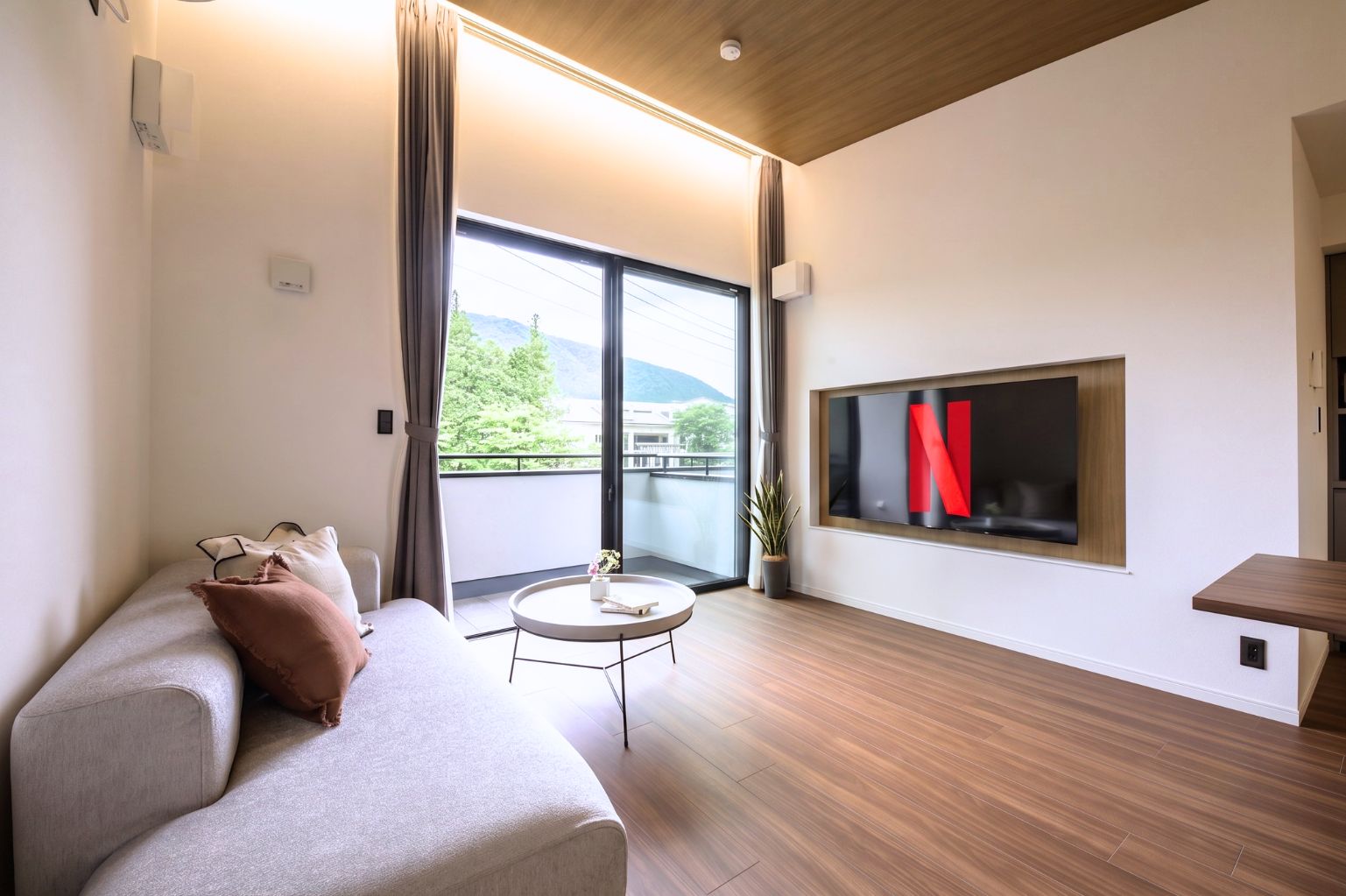 A modern living room with a sofa, a round coffee table, and a large window with a view of greenery and mountains.