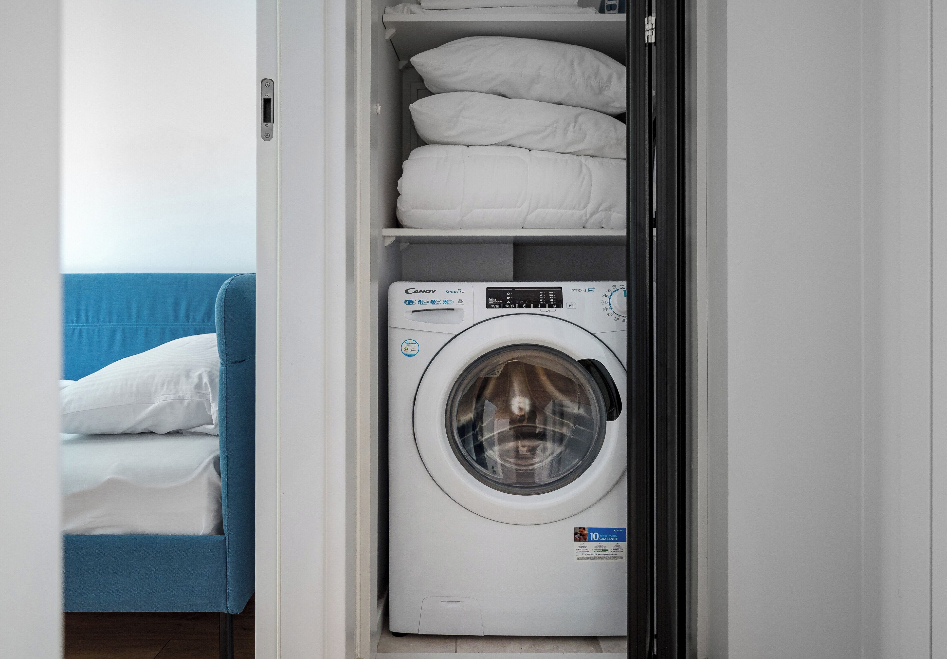 Une pièce de taille modeste équipée d’un lave-linge, d’un lit avec du linge de lit blanc et d’un canapé bleu.