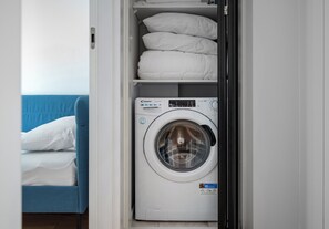 Pieni huone, jossa on pesukone, valkoisilla lakanoiden peitetty sänky ja sininen sohva.