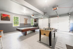 A room with a pool table, a foosball table, and a window with a view of trees.