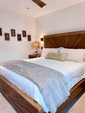 Ein Schlafzimmer mit einem Bettgestell aus Holz, weißer Bettwäsche, einer grauen Tagesdecke, einem Nachttisch mit Lampe und gerahmten Bildern an der Wand.