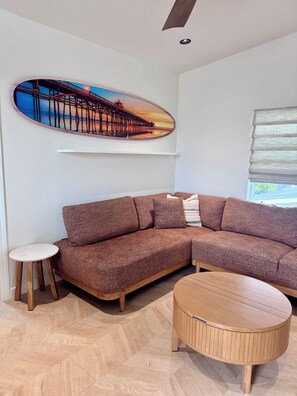 Ein modernes Wohnzimmer mit einem Ecksofa, einem runden Holztisch, einem kleinen Beistelltisch und einem großen Bild im Rahmen an der Wand.