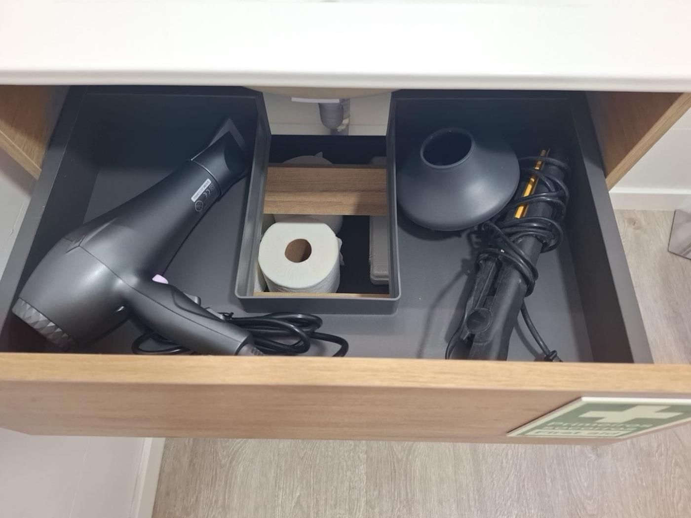 A drawer with a hairdryer, a power cable, a roll of toilet paper, and a black object.