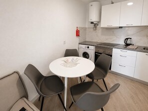 Uma cozinha compacta com máquina de lavar, uma pequena mesa de jantar com cadeiras e um armário branco sobre a pia.