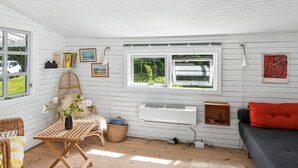 Ein Zimmer mit einem Korbstuhl, einem kleinen Tisch, einem grauen Sofa, einem roten Kissen, einem Fenster mit Blick ins Grüne und einer wandmontierten Klimaanlage.