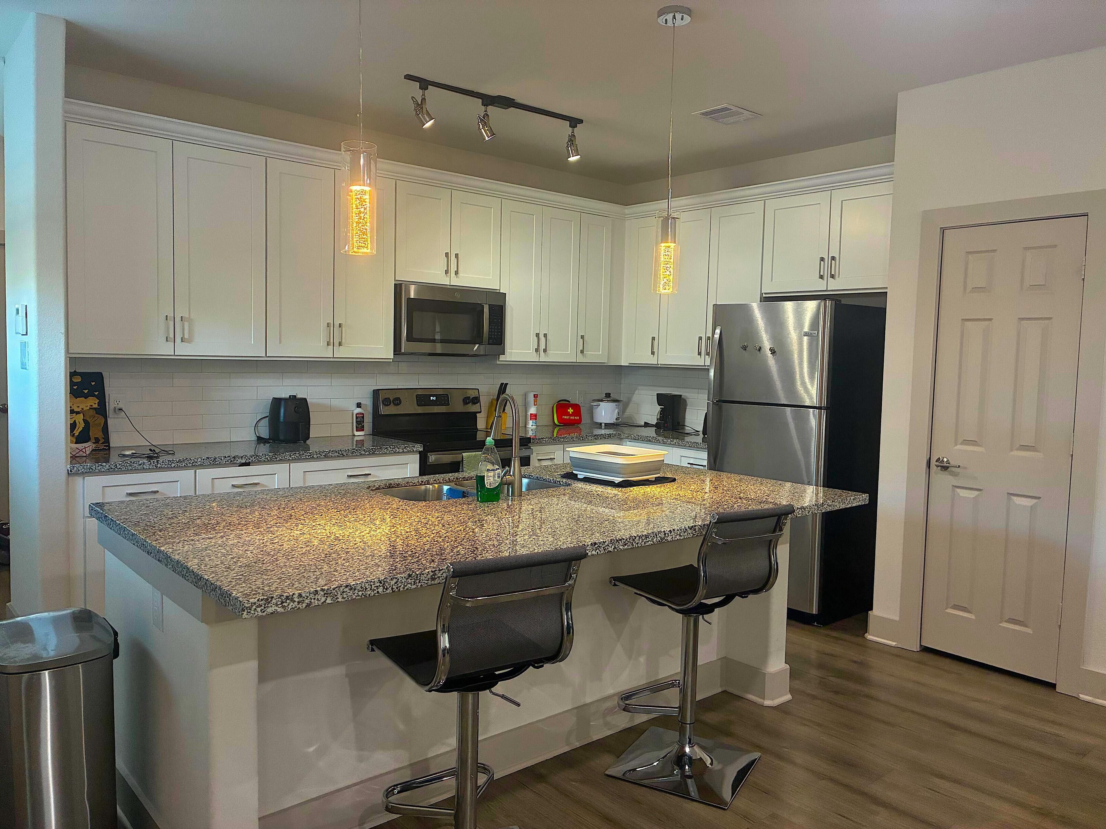 A kitchen with white cabinets, a stainless steel refrigerator, a microwave, a sink, and two bar stools.