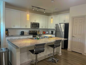 Una cocina con gabinetes blancos, un refrigerador de acero inoxidable, un microondas, un lavamanos y dos taburetes de bar.