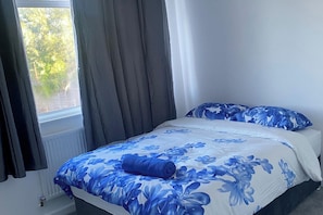 A bed with a floral bedspread, a pillow with a similar design, and a window with curtains. - House Sleeps 7 with Garden and Parking (Erith)