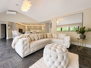 Ein geräumiges Wohnzimmer mit einem großen Ecksofa, einem runden Hocker, einem Essbereich und einer Küche im Hintergrund.