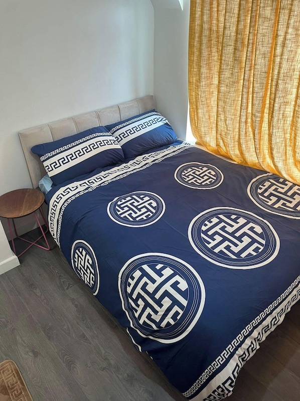A bed with a geometric patterned duvet, a small wooden stool, and a yellow curtain.