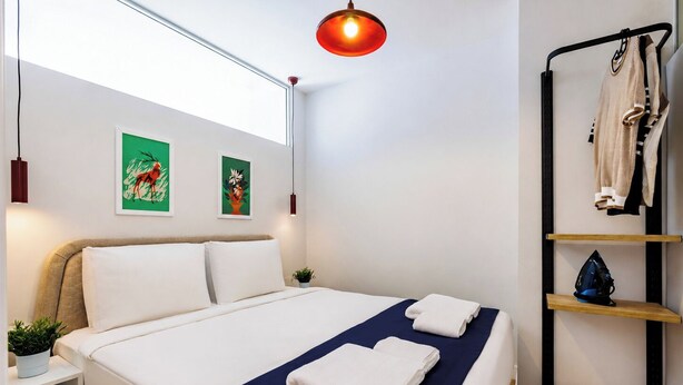 A neatly made bed with white and navy bedding, a small bedside table with a plant, and a wall-mounted shelf with a bag and a decorative item. - Gorgeous 3 Bedroom Apartment At Monnas Suite (Beyoglu)