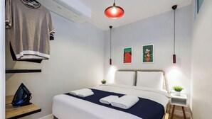 Habitación de hotel con cama, mesitas de noche, perchero, lámpara colgante roja y dos obras de arte en la pared.