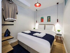 Ein Hotelzimmer mit Bett, Nachttischen, einem aufgehängten Kleidungsstück, einer roten Pendelleuchte und zwei an der Wand befestigten Kunstwerken.