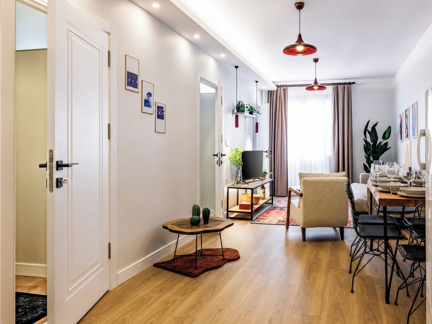 A modern living room with a wooden floor, a TV stand, a sofa, a dining table set for six, and a small side table with a vase.