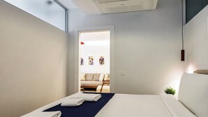Ein modernes Hotelzimmer mit Bett, Handtüchern, einer kleinen Pflanze und Blick auf einen Wohnbereich mit Sofa und Kunstwerken.
