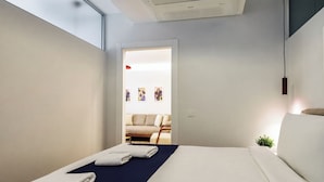 Quarto de hotel moderno com cama, toalhas, uma pequena planta e vista para uma sala de estar com sofá e obras de arte.