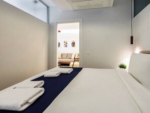 Ein modernes Hotelzimmer mit Bett, Handtüchern, einer kleinen Pflanze und Blick auf einen Wohnbereich mit Sofa und Kunstwerken.