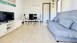 Una sala de estar con televisor, un sofá gris, una mesa de comedor con sillas y una puerta.