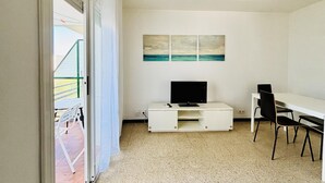Una habitación con televisión, escritorio y silla, una ventana con cortinas y un cuadro en la pared.