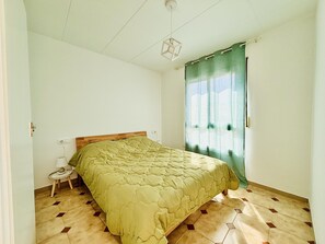 Ein Schlafzimmer mit Bett, Nachttisch, Lampe, einem Fenster mit Vorhängen und einem Boden mit geometrischem Muster.