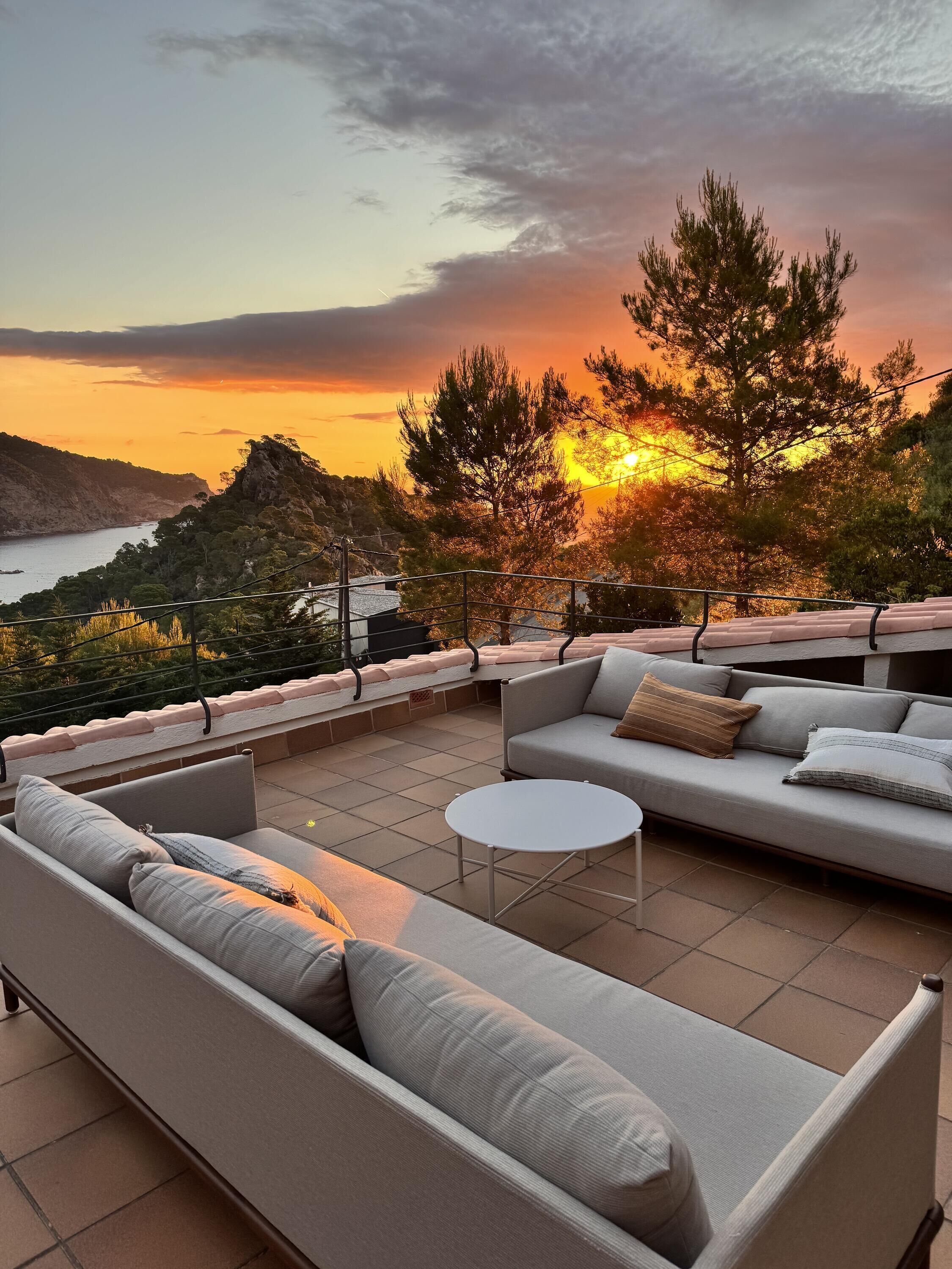 A terrace with a sofa, a small table, and a view of the sunset over a body of water.