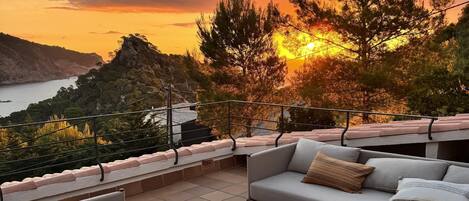 A terrace with a sofa, a small table, and a view of the sunset over a body of water.