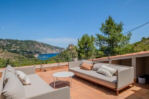 A terrace with a sofa, a small round table, and a view of the sea and hills.