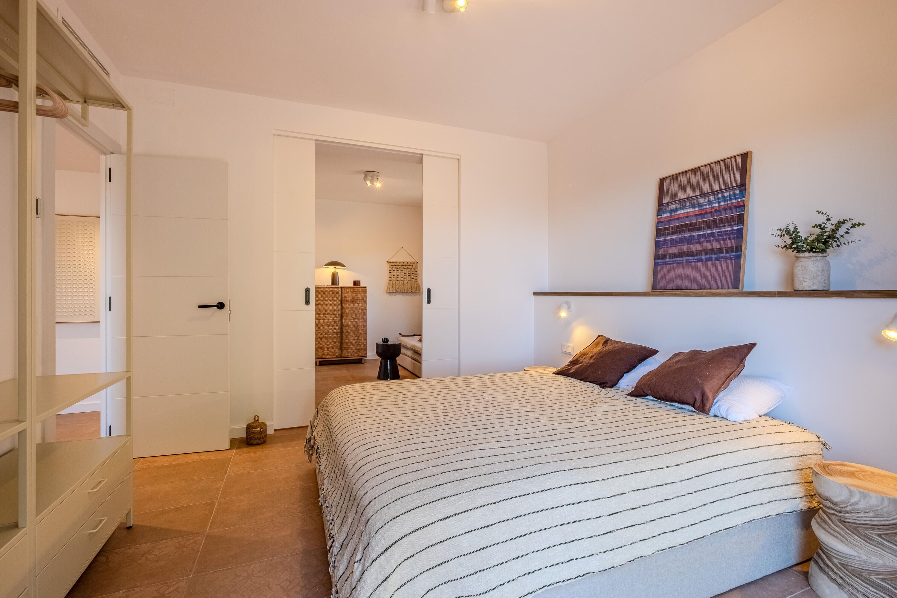 A bedroom with a bed, a wardrobe, a door leading to another room, a wall-mounted shelf, and a wall-mounted light fixture.