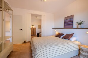 A bedroom with a bed, a wardrobe, a door leading to another room, a wall-mounted shelf, and a wall-mounted light fixture.
