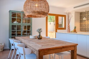 A dining area with a wooden table and white chairs, a wicker pendant light, a green cabinet with glass doors, and a kitchen counter with decorative items.