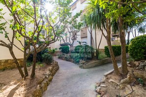A pathway surrounded by trees and plants, leading to a building with a stone wall and a small garden area. - AJ-17 Apartment near the center, large community with pool (Calella de Palafrugell)
