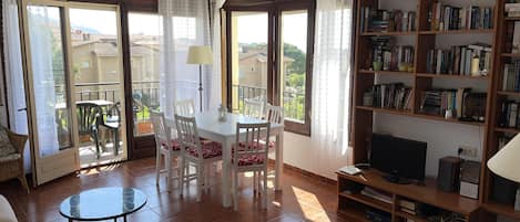 A living room with a dining area, a bookshelf, a TV, and a balcony with a view.