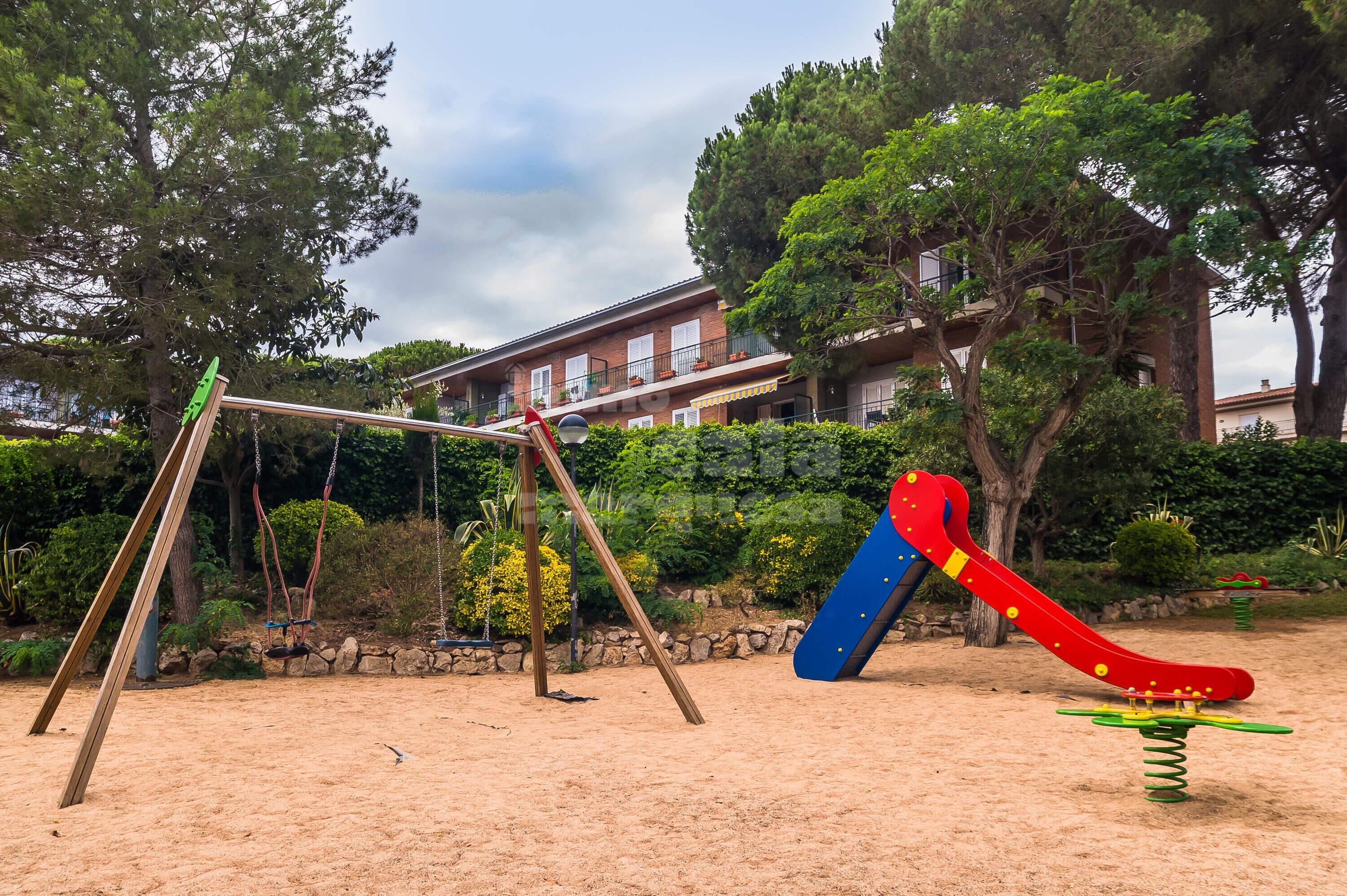 A playground with swings and a slide, surrounded by trees and a building in the background.