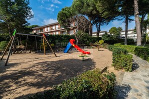 A playground with swings and slides, surrounded by trees and a building in the background. - AJ-17 Apartment near the center, large community with pool (Calella de Palafrugell)