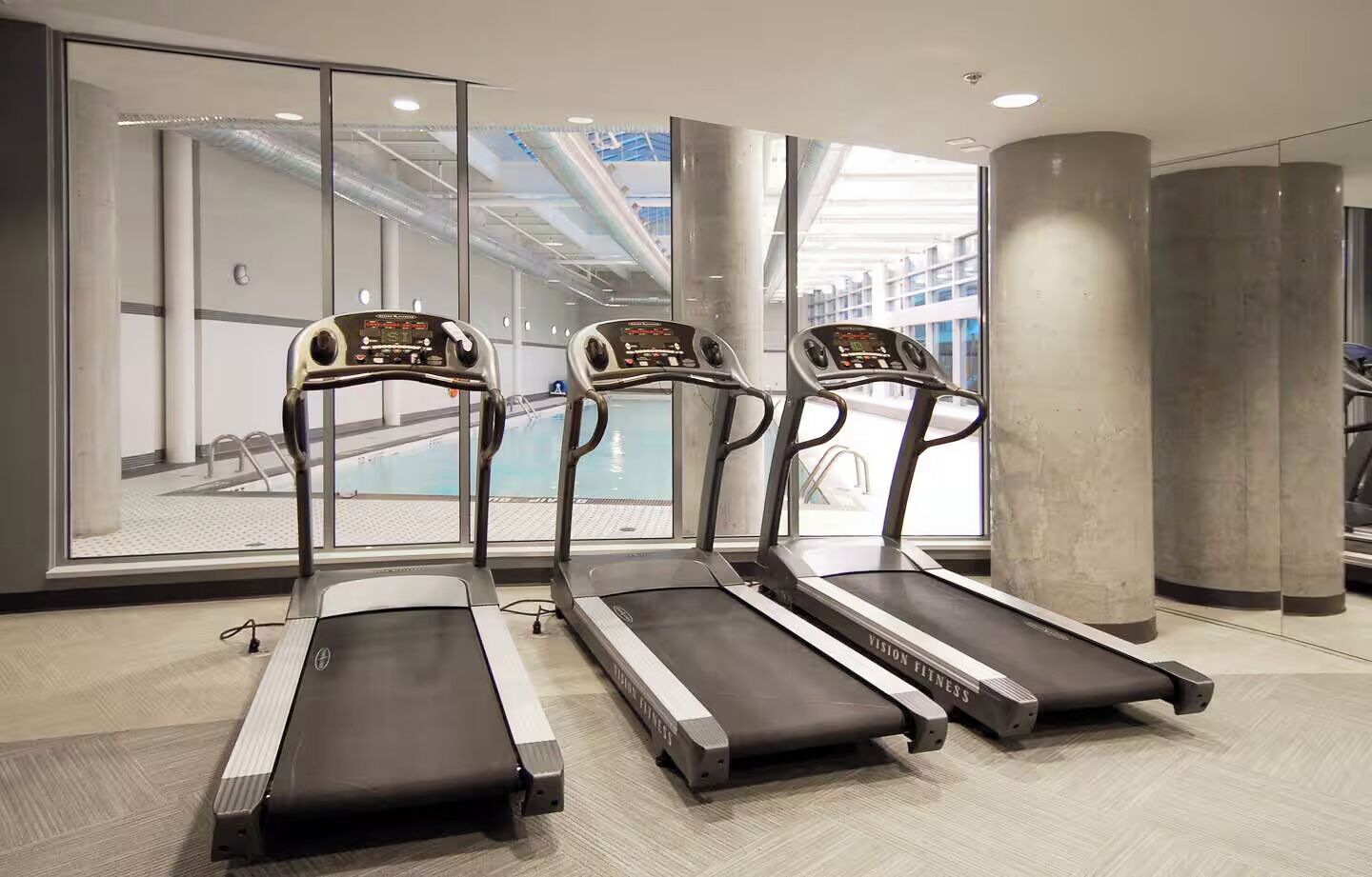 Drei Laufbänder in einem modernen Fitnessstudio mit großen Fenstern und Blick auf ein Schwimmbecken.