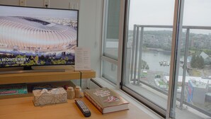 Un televisor mostrando un estadio, un libro titulado 'The Ultimate Sneaker Book' y un control remoto sobre una mesa de madera junto a una ventana con vistas a la ciudad.